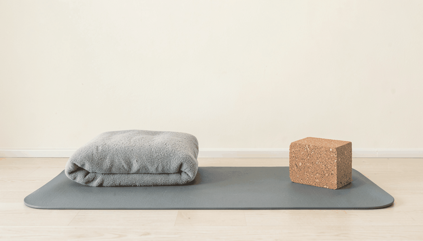 A neatly arranged yoga mat with a folded blanket and cork block, set against a light cream wall, serene and minimal