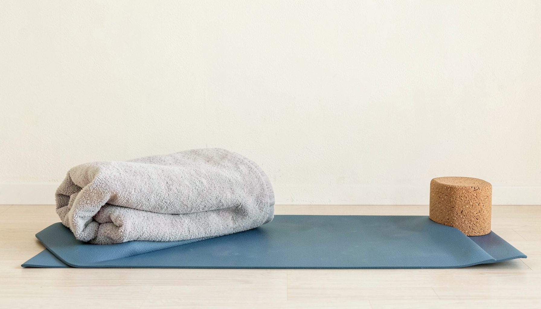 A neatly arranged yoga mat with a folded blanket and a cork block beside it, set against a light cream wall, evoking a quiet mood