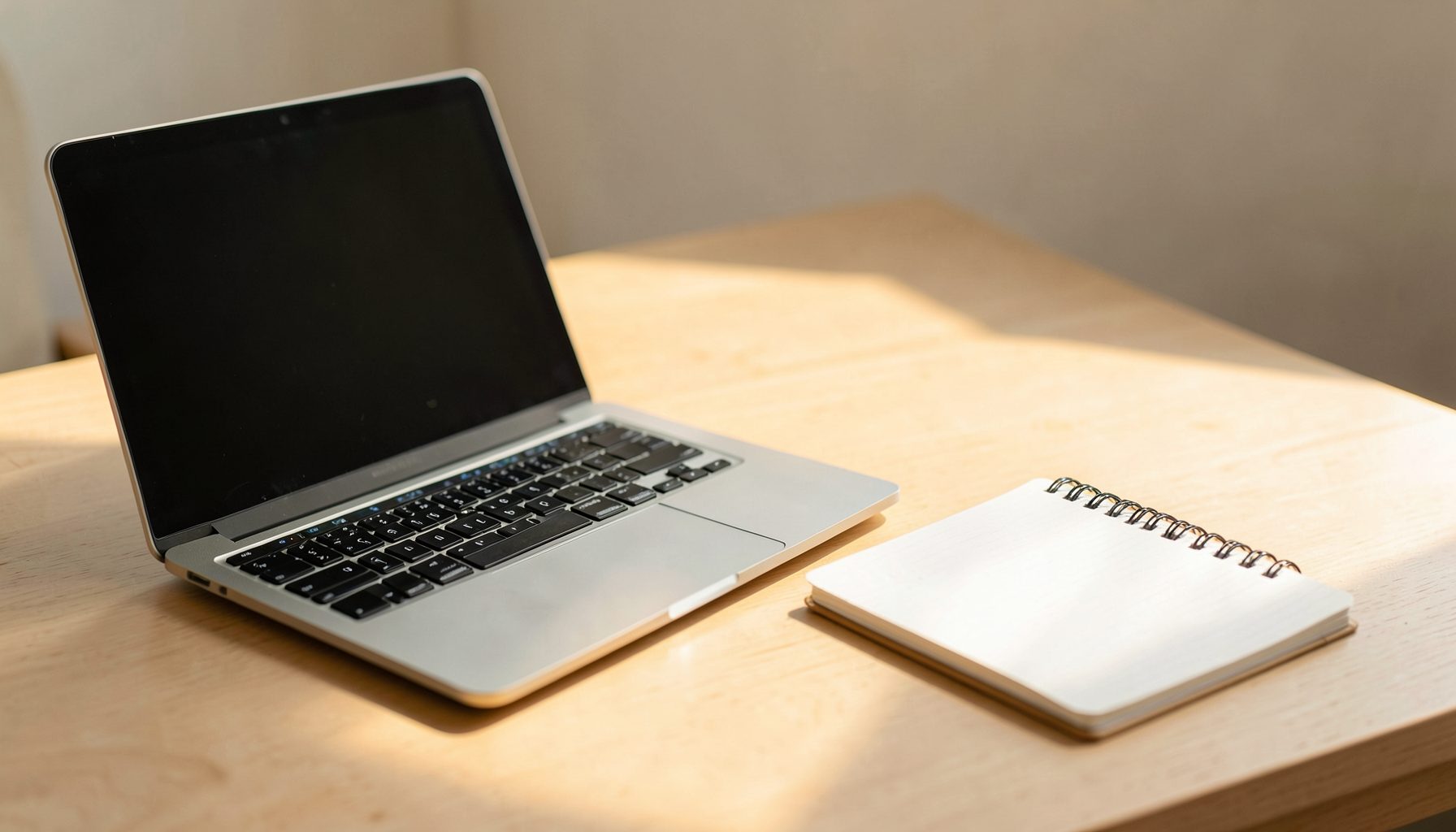 A simple, uncluttered workspace with a laptop and a notebook on a light wood desk, bathed in warm afternoon light, suggesting quiet focus