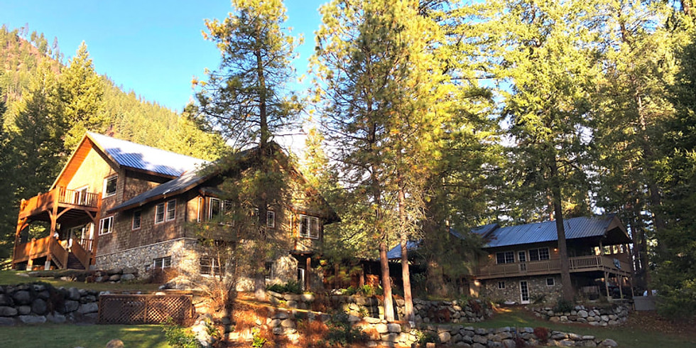 A large wooden lodge-style building surrounded by tall pine trees and stone landscaping under a clear blue sky, tranquil